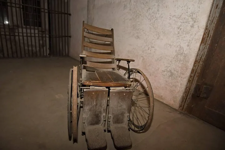 Old wooden wheelchair sitting in a sparse, dimly lit room with bars.